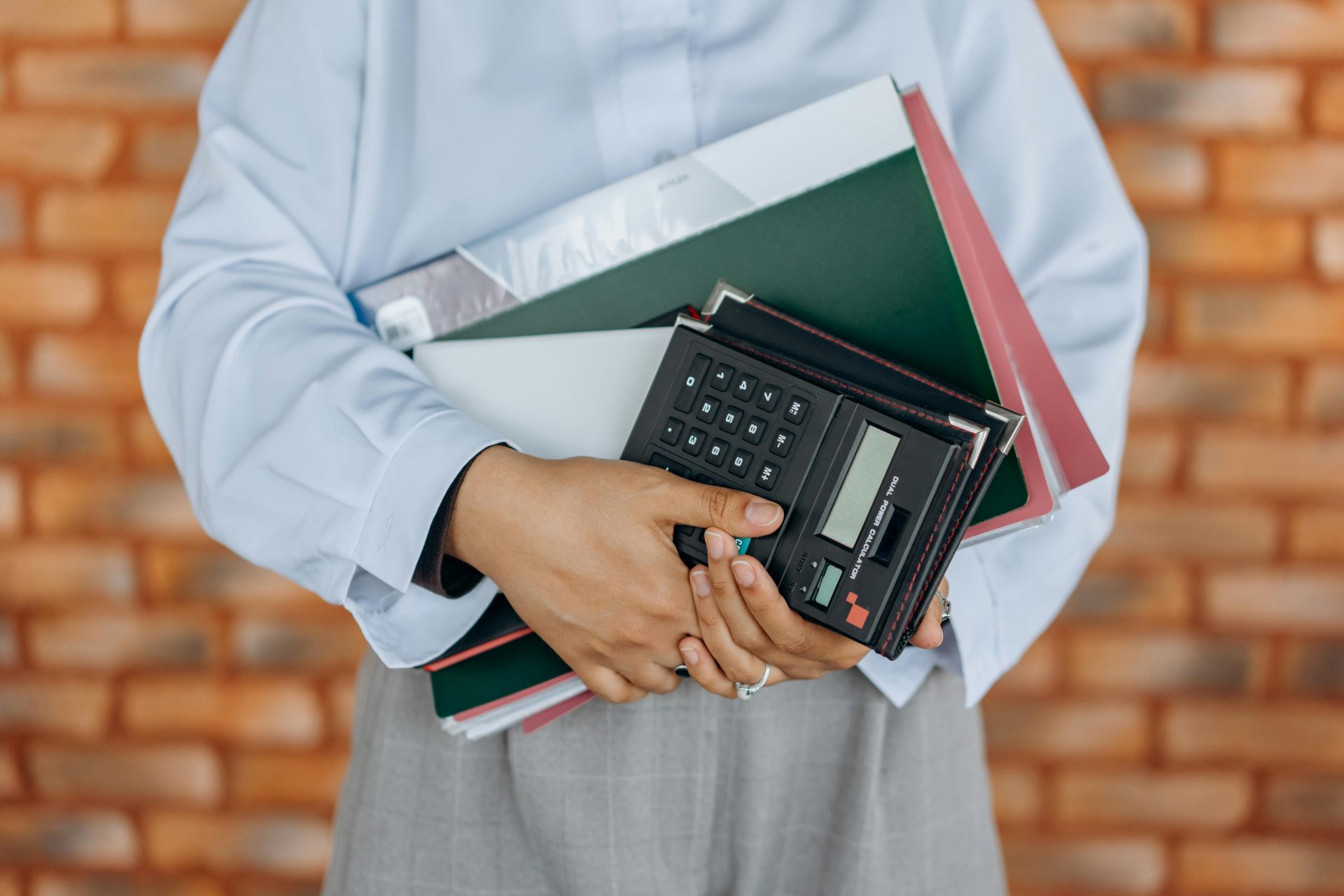 Gros plan sur une personne tenant une calculatrice et des dossiers devant un mur de briques.